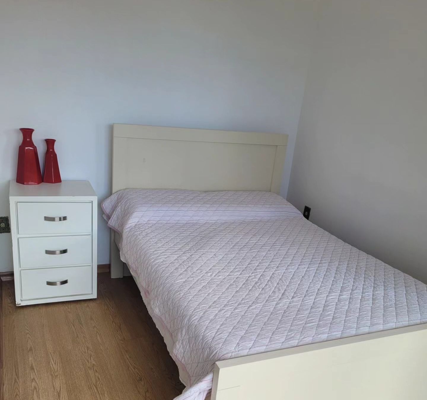 Suíte com cama arrumada, criado-mudo branco e piso de madeira.